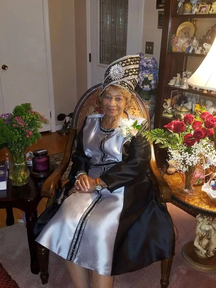 Missionary Katherine Cash Griffey seated in an armchair, wearing a silver-and-black satin robe and a matching wide-brimmed beaded hat, surrounded by flowers.